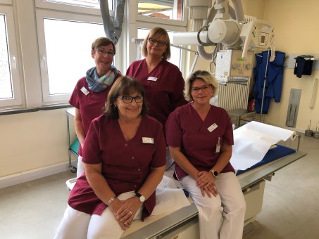 Vier Damen mit rotem Kasak sitzen auf einer Behandlungsliege im Röntgenraum