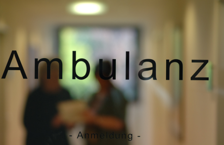 Den Gang zur Ambulanz durch eine Glastür verschwommen zu sehen. Im Vordergrund der Schriftzug Ambulanz, klein darunter Anmeldung.
